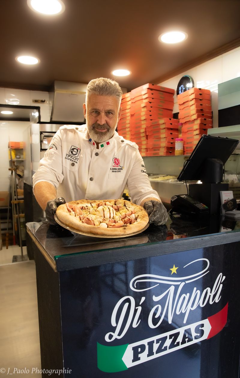 Rémy in weißer Jacke zeigt eine Pizza hinter der Theke „Di Napoli Pizzas“.