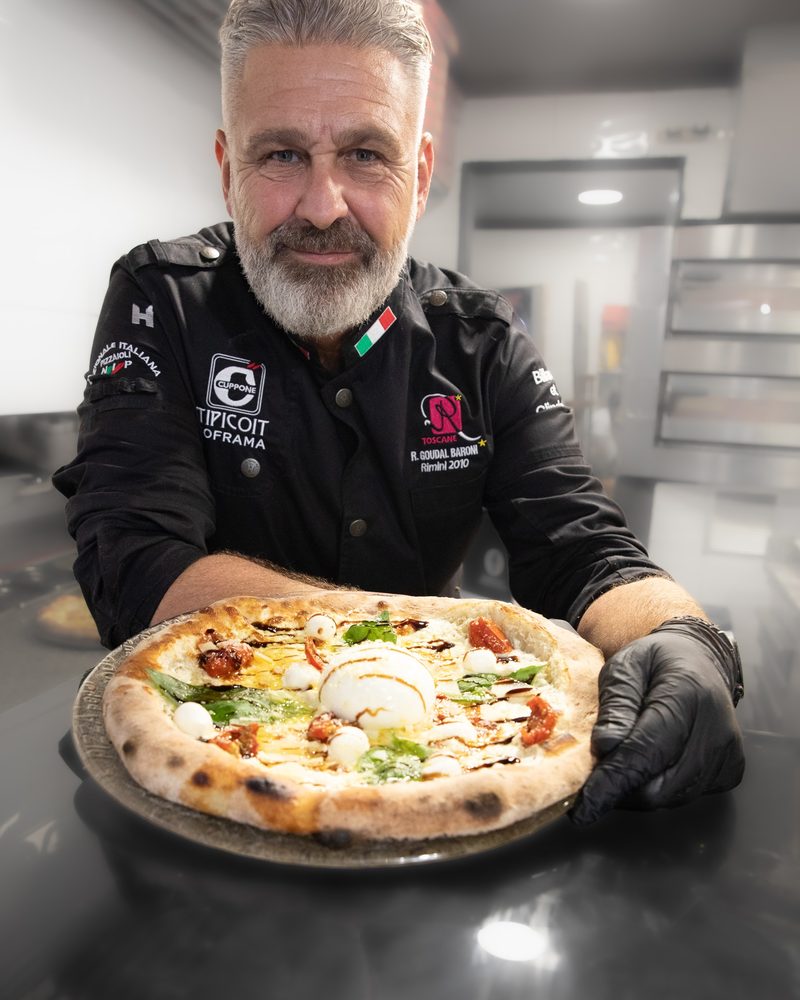 Rémy, Pizzabäcker in schwarzer Jacke, zeigt eine neapolitanische Pizza mit Burrata vor dem Ofen.